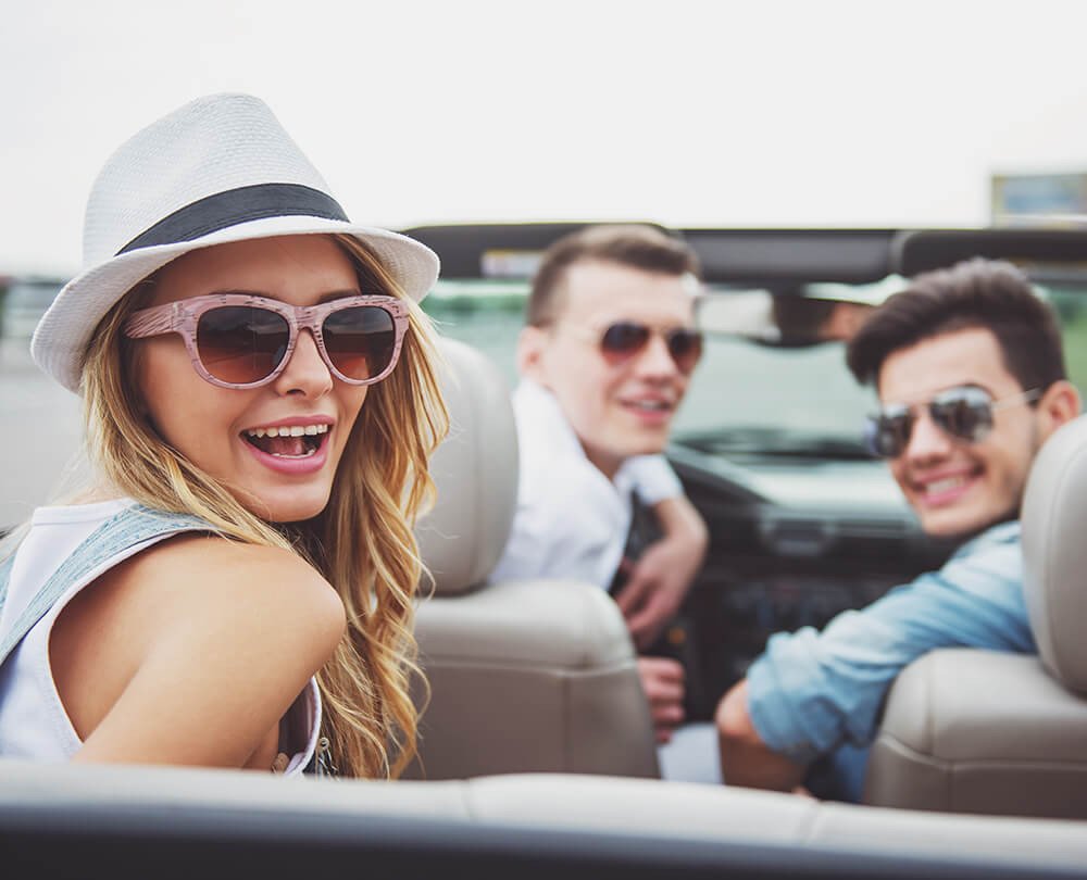 dentro de um carro conversível uma moça no banco de tras e dois rapazess no banco da frente todos sorriem