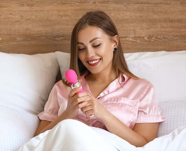 imagem mulher jovem sentada na cama segurando e admirando vibrador varinha mágica cor pink