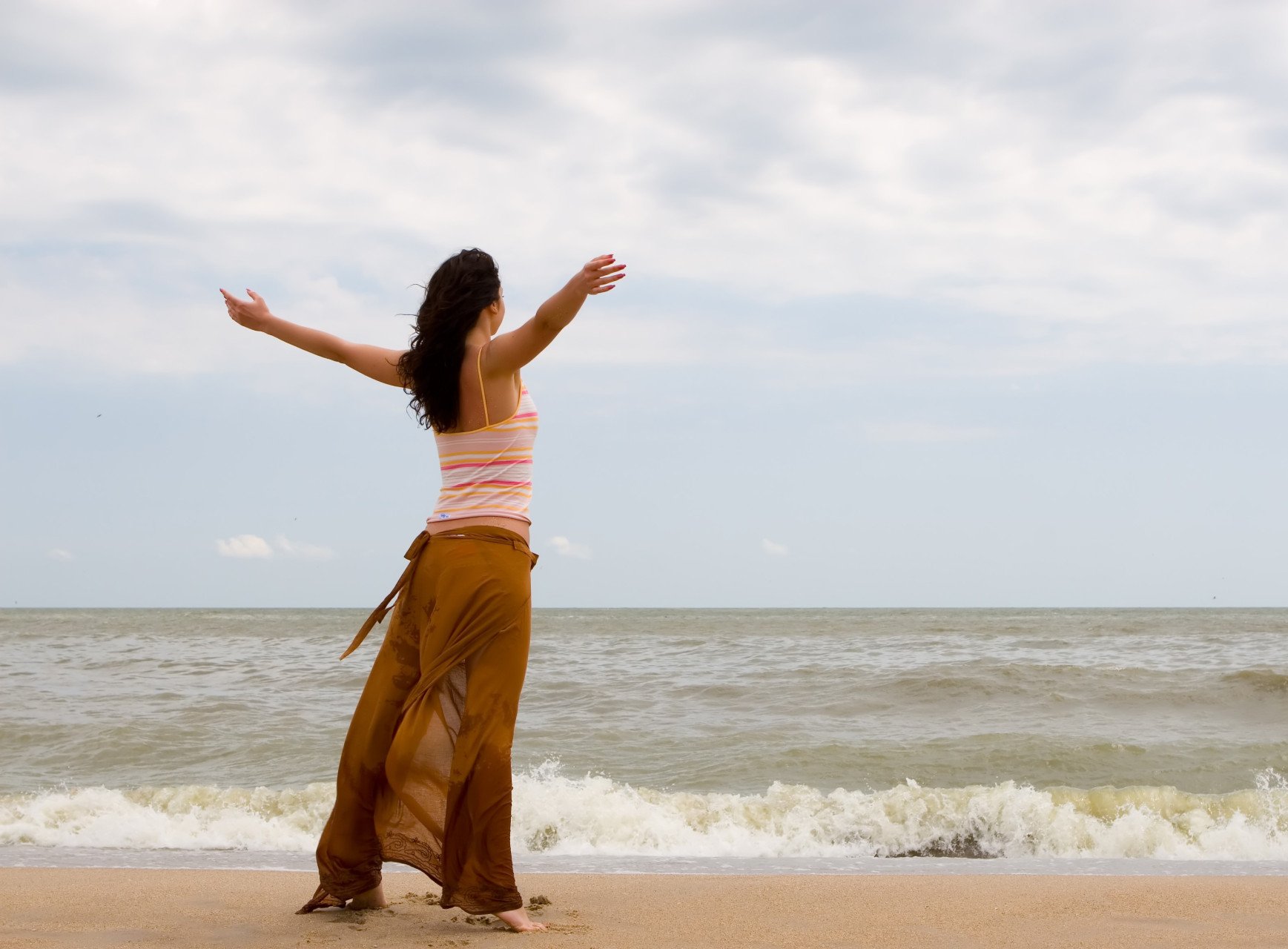 Foto de uma mulher de costas na praia olhando o mar com os braços abertos